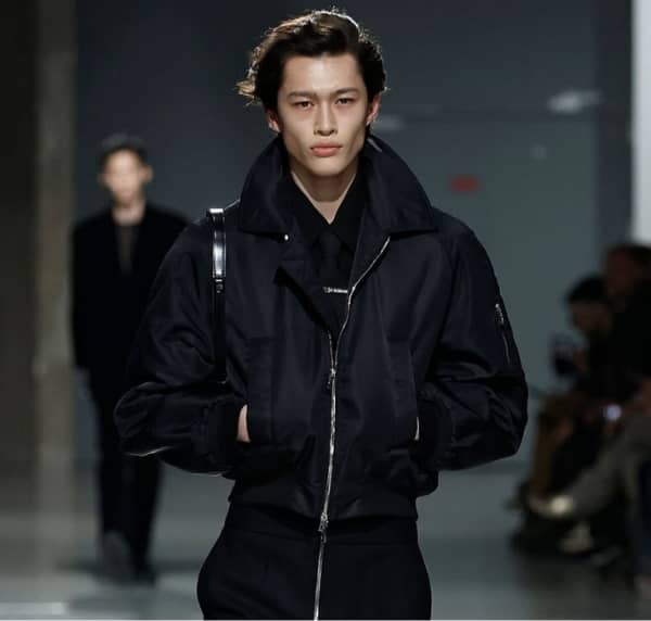 Male model walking runway in a black designer bomber jacket by LGN Louis Gabriel Nouchi, featuring a high collar and minimalist tailoring during a fashion show.