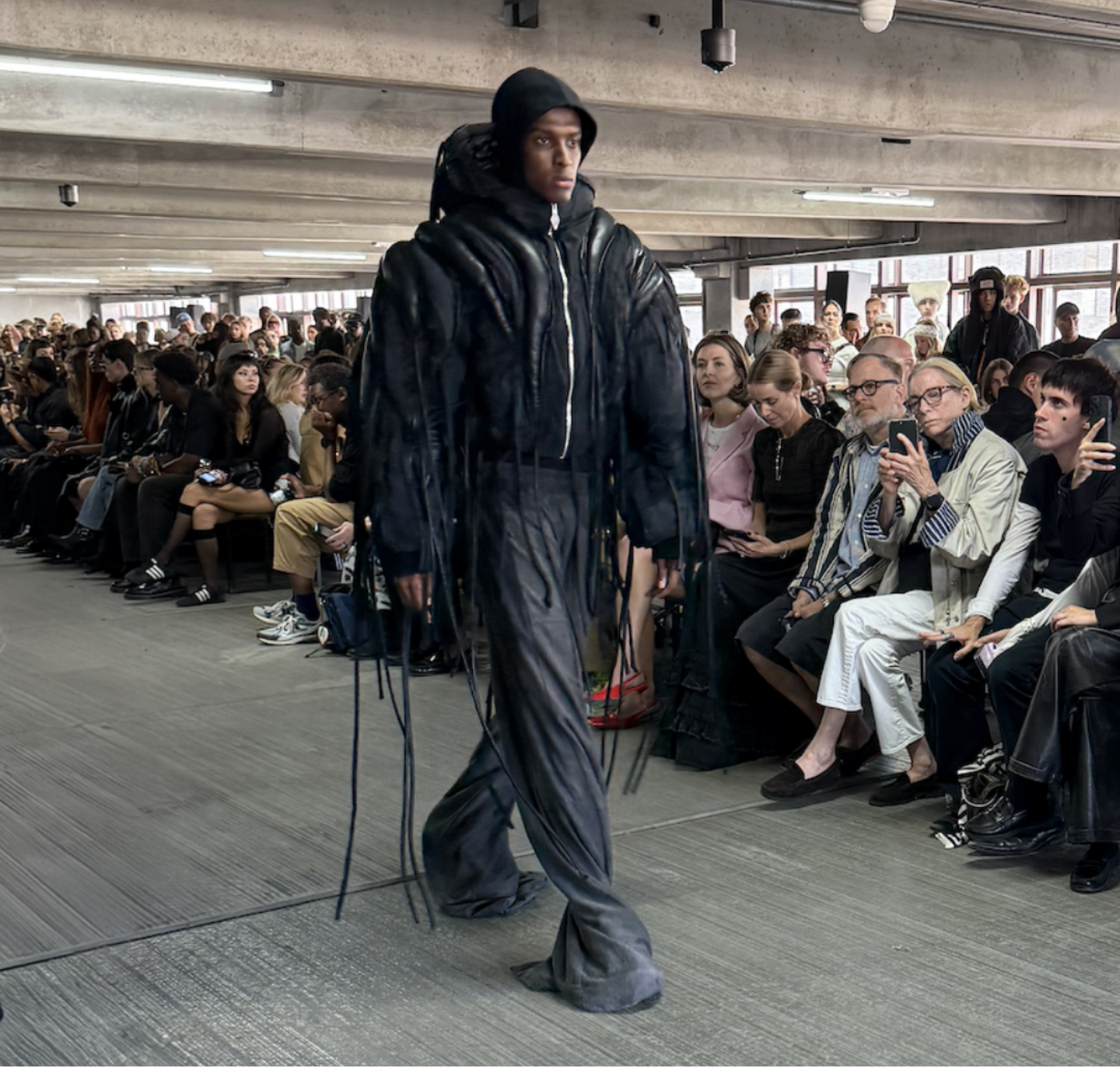 Model walking the runway in a black sculptural hooded jacket with elongated fringe detailing at the Han Kjøbenhavn show, surrounded by a full seated audience in an industrial venue.