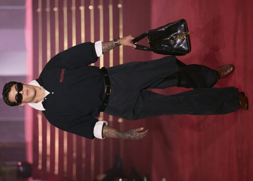 Model walks the runway in an oversized navy polo, wide-leg trousers, brown leather shoes, and black sunglasses, carrying a large black leather tote under warm pink lighting.