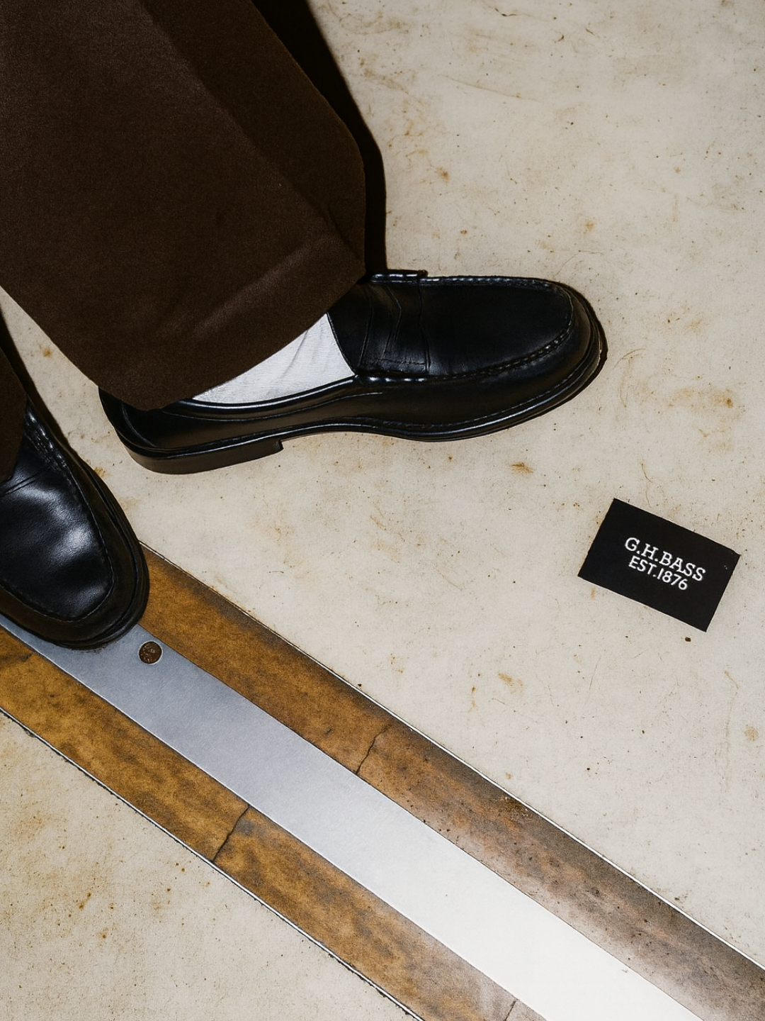 Close-up of a person wearing black G.H. Bass penny loafers with white socks and brown trousers, standing on a textured floor beside a small G.H. Bass logo card reading “EST. 1876.”