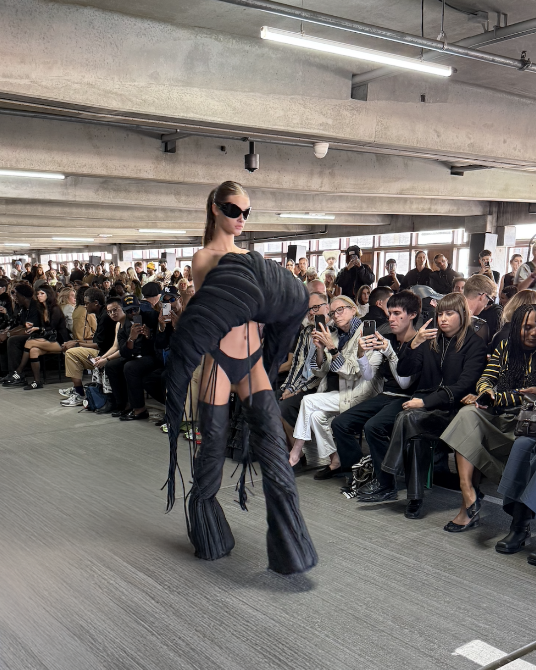 Han Kjøbenhavn runway — model in sculptural black cut-out look with oversized textured sleeves, thigh-high boots, and futuristic sunglasses, embodying Scandinavian avant-garde fashion.
