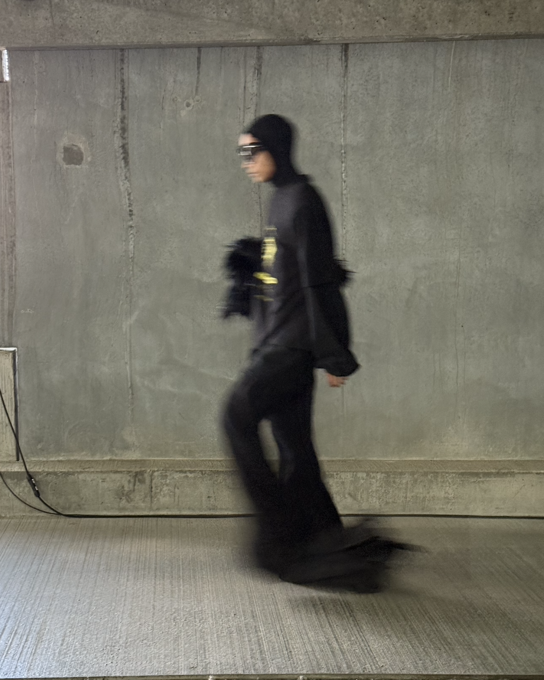 Han Kjøbenhavn runway look in all-black — sculptural streetwear silhouette with oversized hood, futuristic sunglasses, and fluid tailoring against brutalist concrete backdrop.