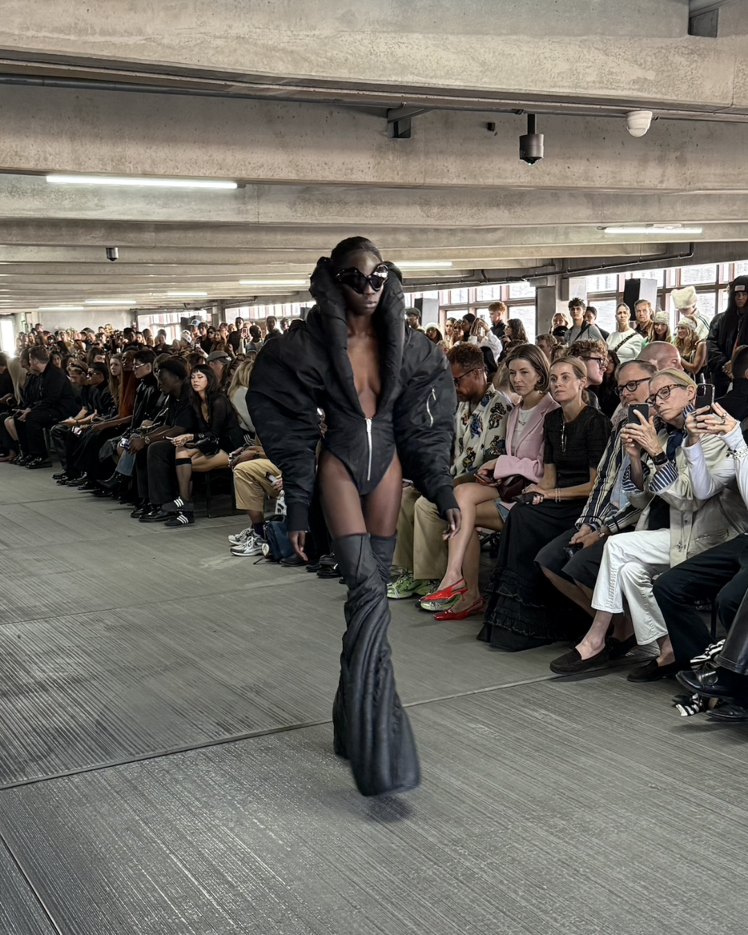 Han Kjøbenhavn runway — model in sculptural black puffer bodysuit with exaggerated sleeves, thigh-high boots, and oversized sunglasses, embodying Scandinavian avant-garde fashion.