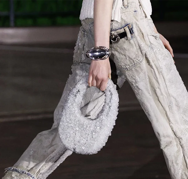 Close-up of a model carrying a crystal-embellished Coperni Swipe Bag while wearing distressed textured jeans and a reflective sculptural bracelet during a fashion runway presentation.