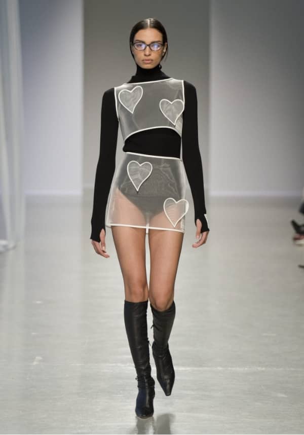 Model walking the runway in a sheer white heart-panel dress layered over a black turtleneck bodysuit with knee-high boots at the FW25 Kimhekim show.