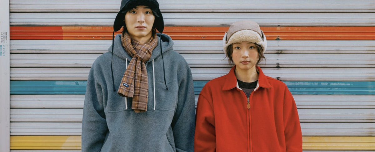 Two models wearing oversized Kappy Design outerwear, featuring a grey hoodie with a plaid scarf and black earflap hat, and a red zip-up jacket with a shearling-trimmed hat, against a colourful striped metal shutter.