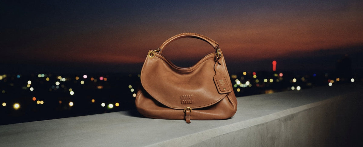 Miu Miu brown leather handbag with signature logo set against an evening cityscape – shop Miu Miu bags at ELLIS ROSCH