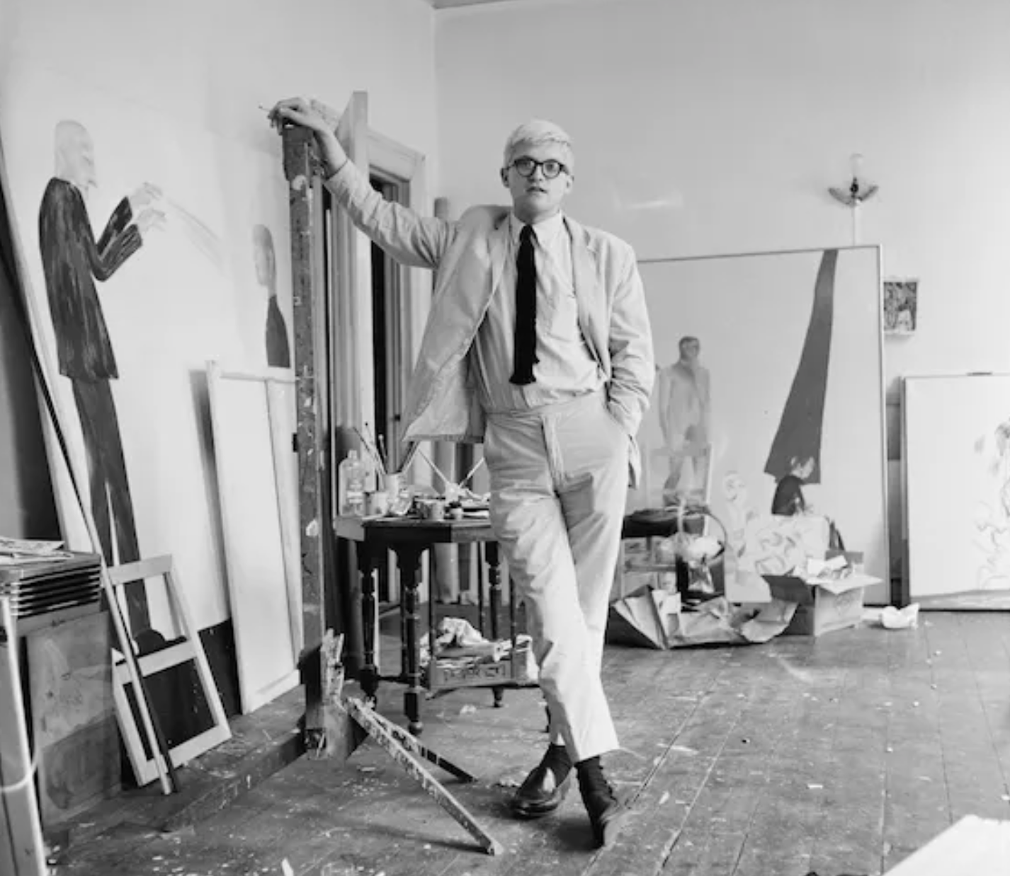 Black-and-white photograph of a David Hockney in a tailored suit and penny loafers standing in an artist’s studio surrounded by large canvases and painting tools, embodying mid-century creative style.