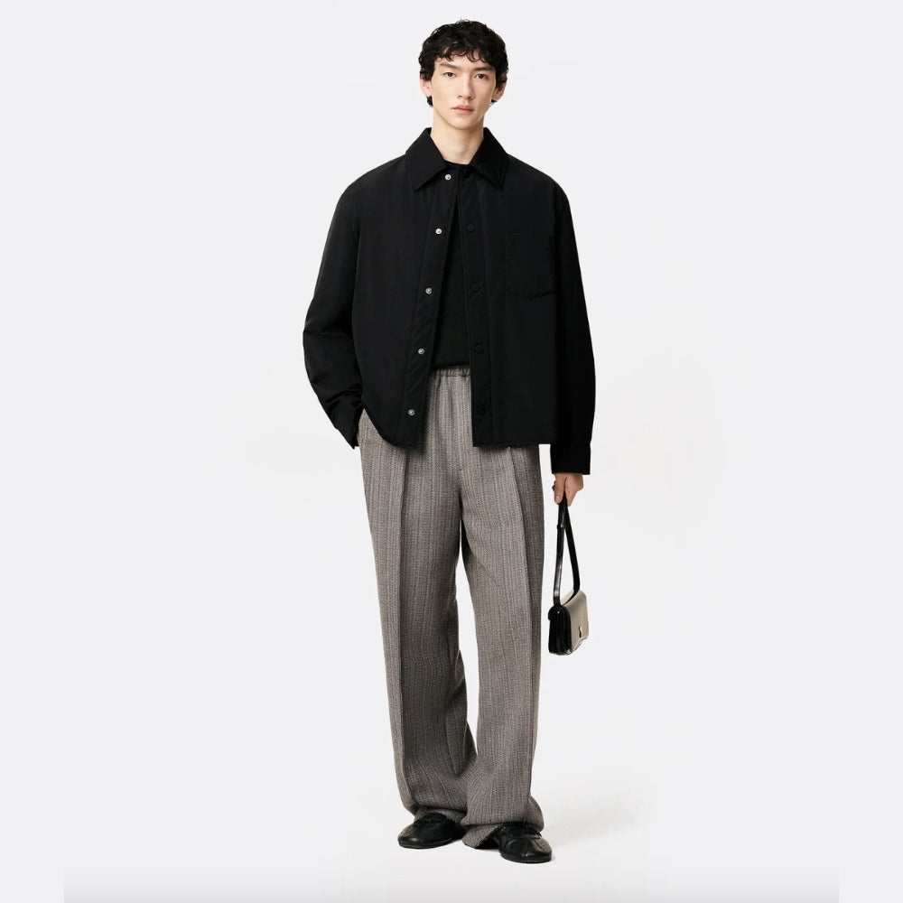 Model wearing a black button-up jacket over a black top with relaxed-fit grey textured trousers, styled with black leather shoes and a small structured shoulder bag, presented against a clean neutral background for ELLIS ROSCH menswear.