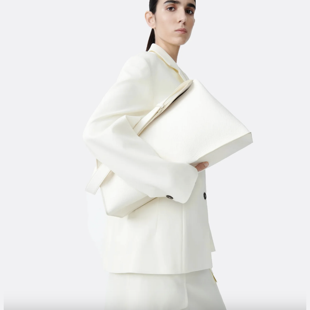 Model wearing a tailored off-white blazer and matching trousers, carrying an oversized structured white leather tote bag under the arm, styled against a clean neutral background.