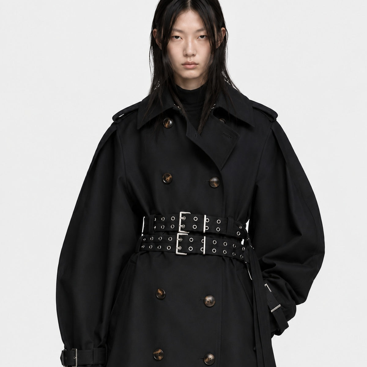 Editorial model wearing aoversizedblack designer trench coat with a sculptural gold hand detail and black leather skirt.