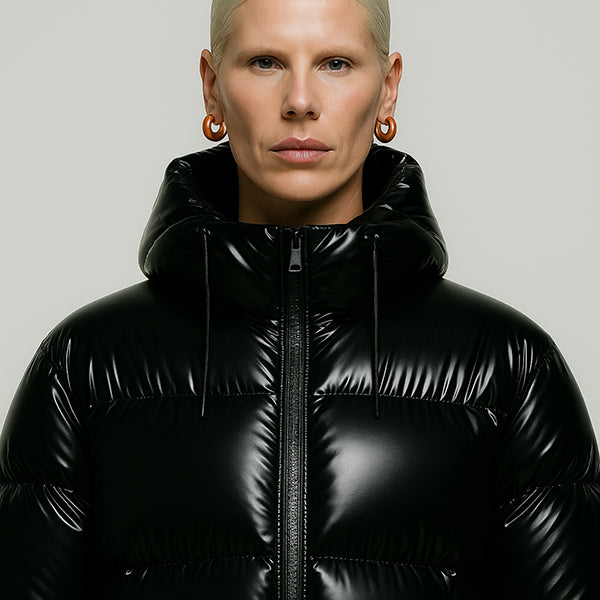 Feminine Model wearing a premium black glossy puffer jacket with hood curated from ELLIS ROSCH — minimalist winter outerwear in modern sculptural silhouette.
