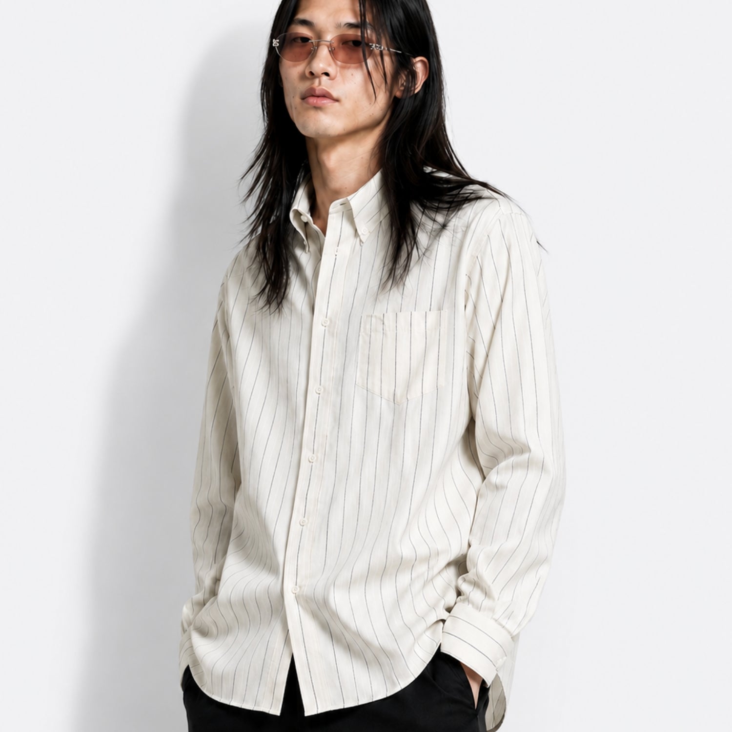 Model wearing a relaxed off-white striped button-up shirt with a pointed collar and chest pocket. Styled with dark tailored trousers and tinted narrow sunglasses, the look balances understated tailoring with a contemporary, gender-fluid aesthetic and effortless minimalist styling.