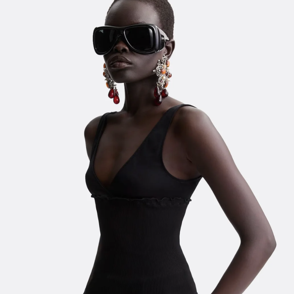 Model wearing a fitted black ribbed sleeveless dress with a deep V neckline, styled with oversized black visor sunglasses and statement chandelier earrings with red and orange accents, against a neutral background.
