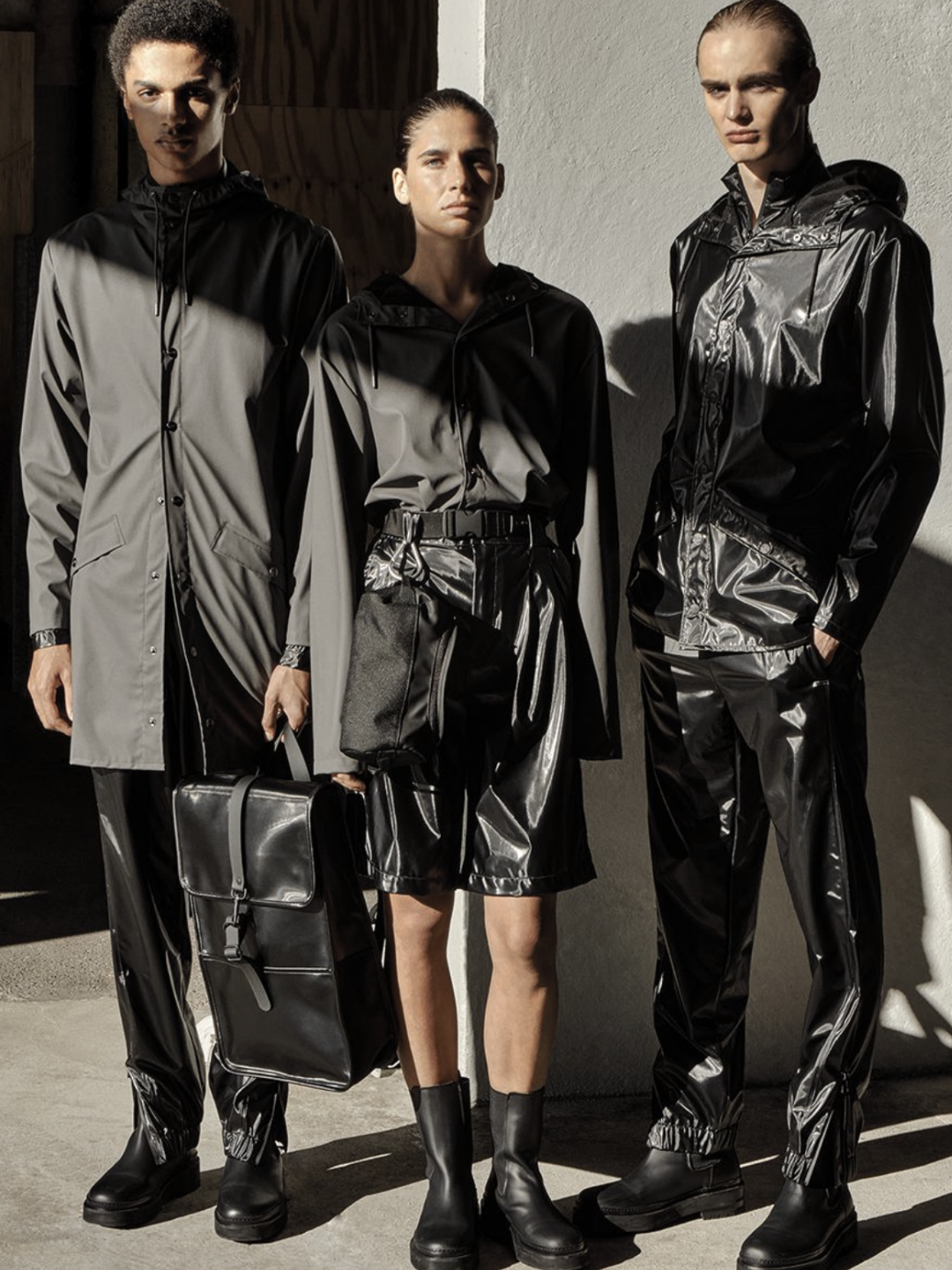 RAINS collection in monochrome — models styled in waterproof raincoats, glossy rainwear trousers, and minimalist boots with structured black backpack and crossbody bag, showcasing Scandinavian functional fashion.